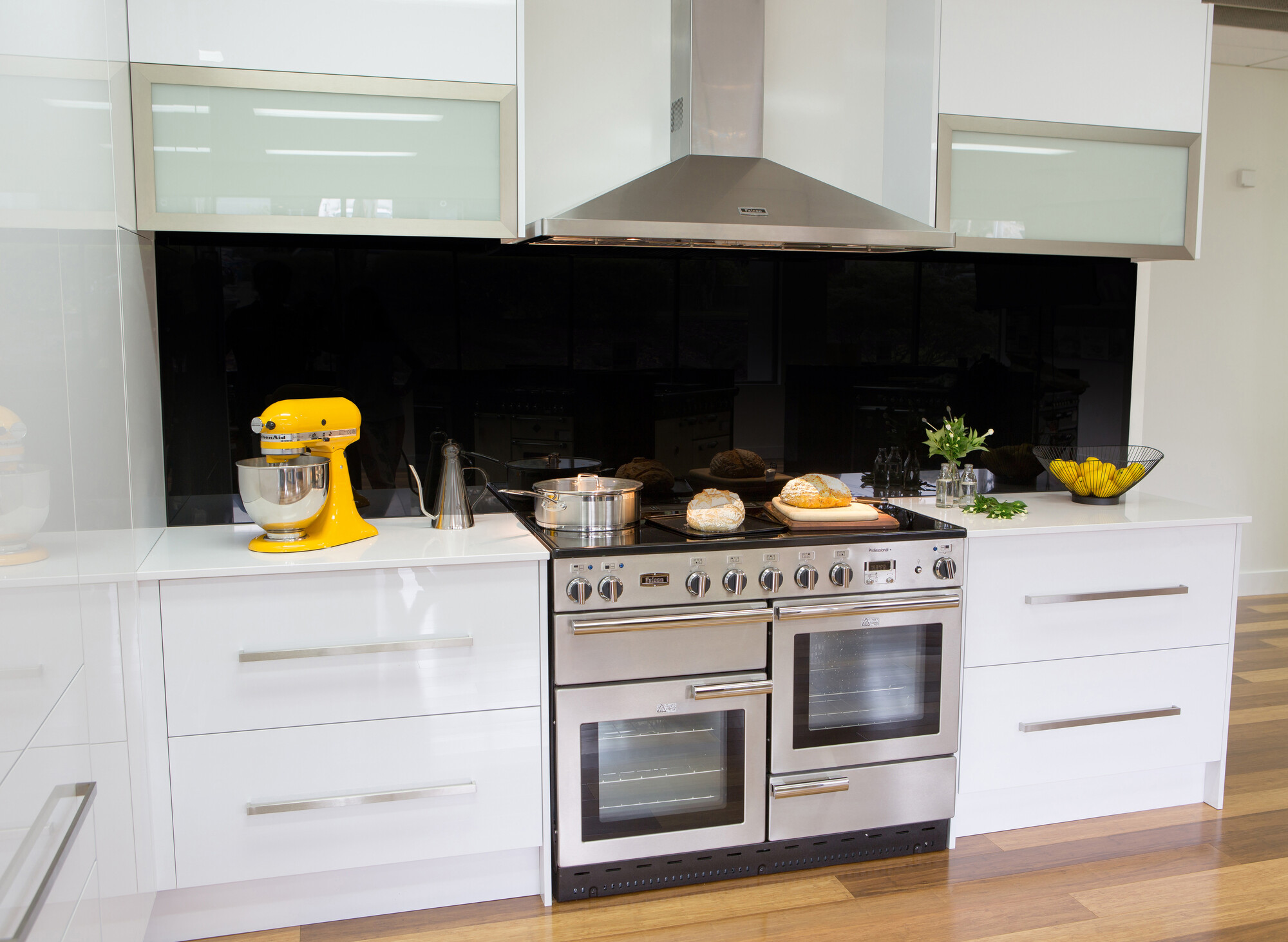 Helle, moderne Küche mit großem Edelstahlherd, gelber Küchenmaschine, Brotlaiben, Topf und frischen Kräutern auf der Arbeitsfläche