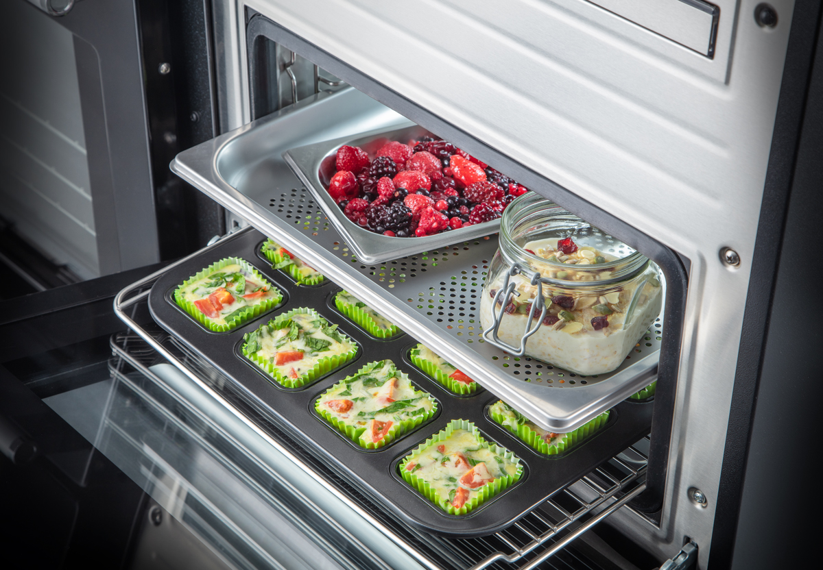 Ein moderner Backofen mit Muffinformen mit Gemüse, einem Glas mit Haferflocken und Nüssen sowie einem Blech mit frischen Beeren.