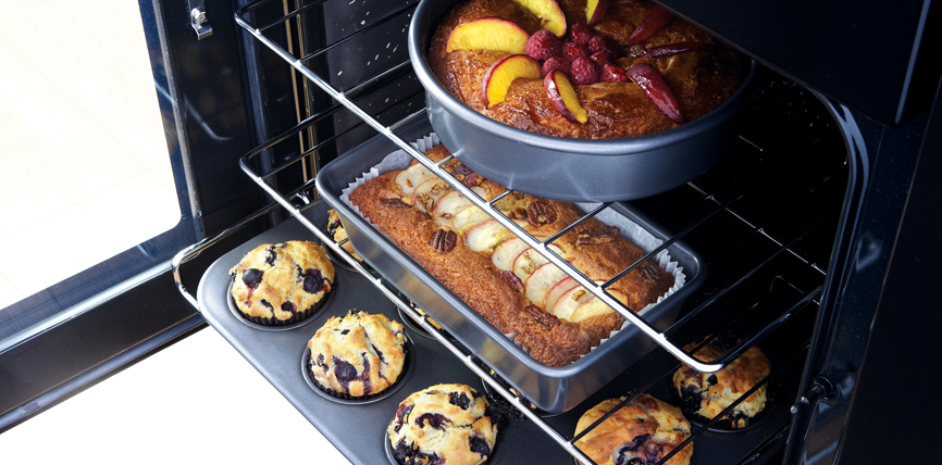 Offener Backofen mit einer runden Kuchenform, einem Bananenbrot und fünf Heidelbeermuffins beim Backen
