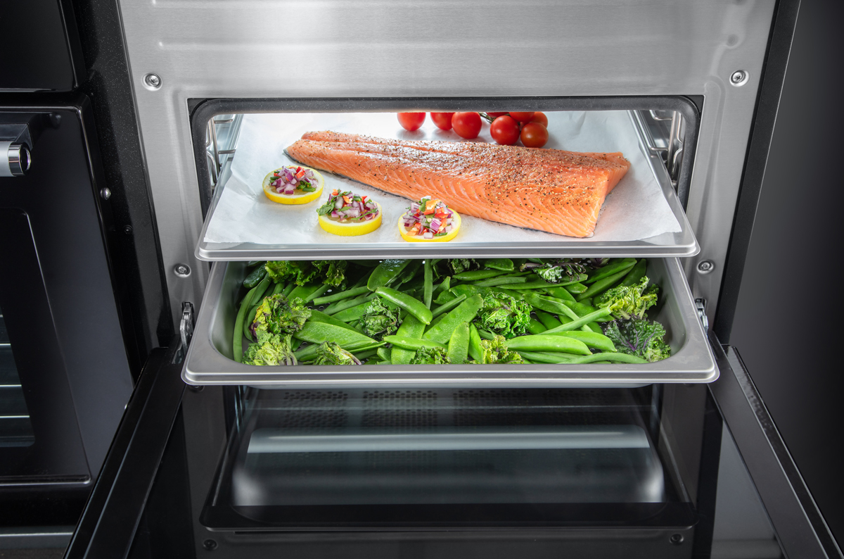 Im geöffneten Ofen garen ein mariniertes Lachsfilet, Zitronenscheiben mit Kräutern und Kirschtomaten auf einem Blech sowie frisches grünes Gemüse auf einer zweiten Ebene.