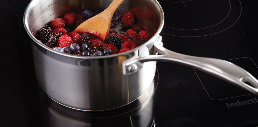 Topf aus Edelstahl mit Himbeeren, Brombeeren und Heidelbeeren auf einem Induktionskochfeld, Holzlöffel rührt Obst