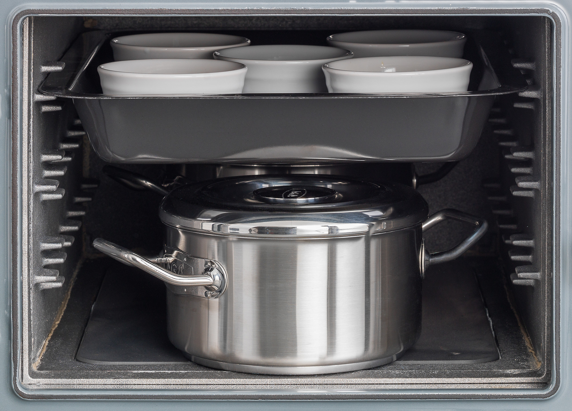 Ein geschlossener Edelstahltopf und mehrere weiße Keramikschalen in einer Backform stehen zusammen in einem Backofen.