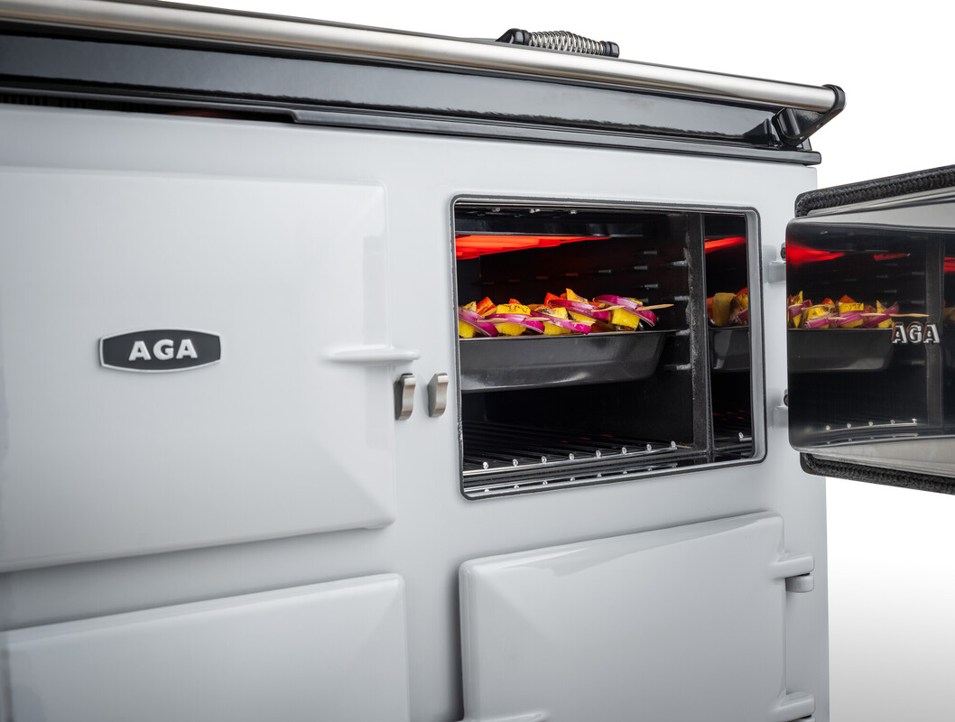 Weißer AGA Küchenherd mit geöffneter Backofentür. Im Ofen befindet sich ein Backblech mit geschnittenem Gemüse, das gebacken wird.