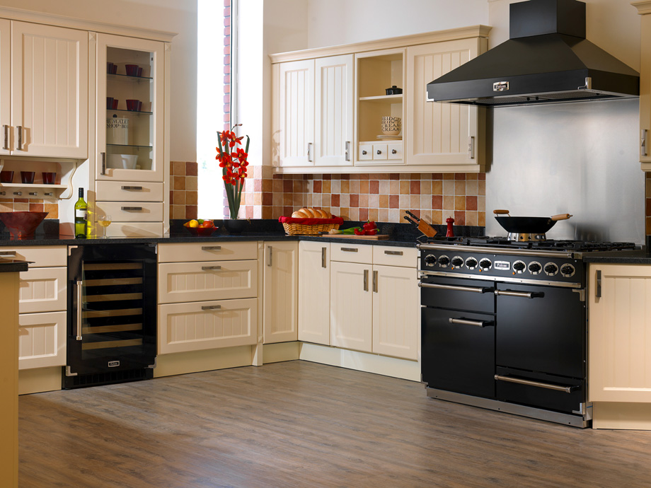 Große, helle Küche im Landhausstil mit cremefarbenen Einbauschränken, schwarzem Herd, Weinkühlschrank, Fliesenspiegel und dekorativen Elementen wie einer Vase mit roten Blumen und frischem Brot auf der Arbeitsplatte