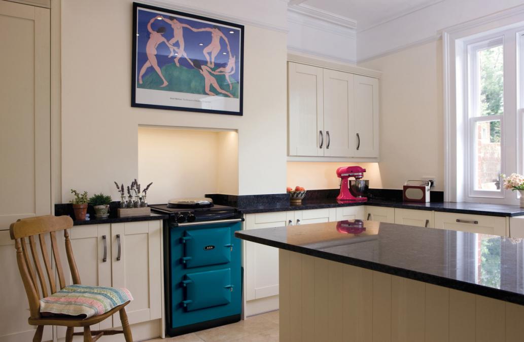 Helle Landhausküche mit cremefarbenen Schränken, schwarzer Granitplatte, türkisem Herd, Holzstuhl und pinker Küchenmaschine