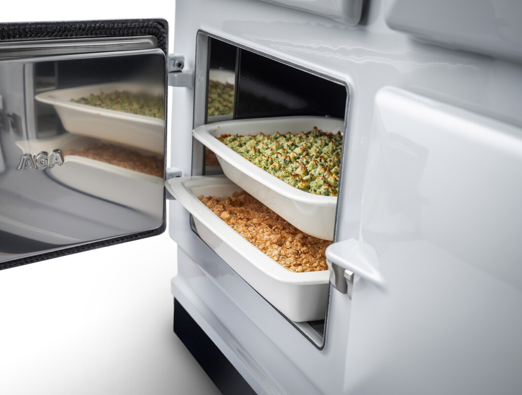 Offener AGA-Backofen mit zwei weißen Keramikformen, gefüllt mit überbackenem Gemüse und Auflauf, moderne Küchenausstattung
