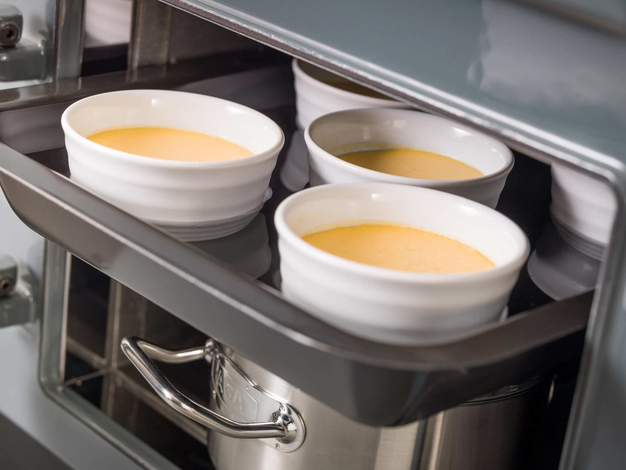Mehrere weiße Keramikförmchen gefüllt mit gelber Creme stehen auf einem Backblech im geöffneten Ofen.