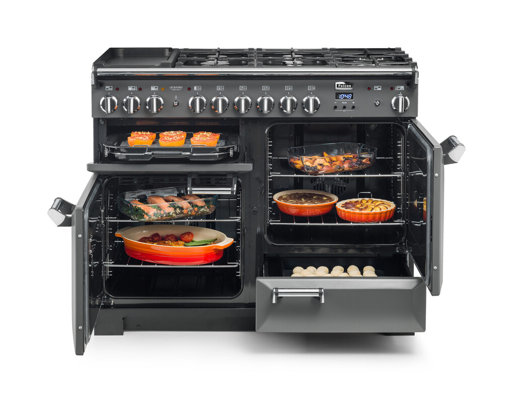 Moderner schwarzer Standherd mit Gasherd und mehreren geöffneten Backöfen, die mit verschiedenen Gerichten wie gefüllten Paprika, gebackenem Gemüse, Fleisch und Kuchen gefüllt sind.