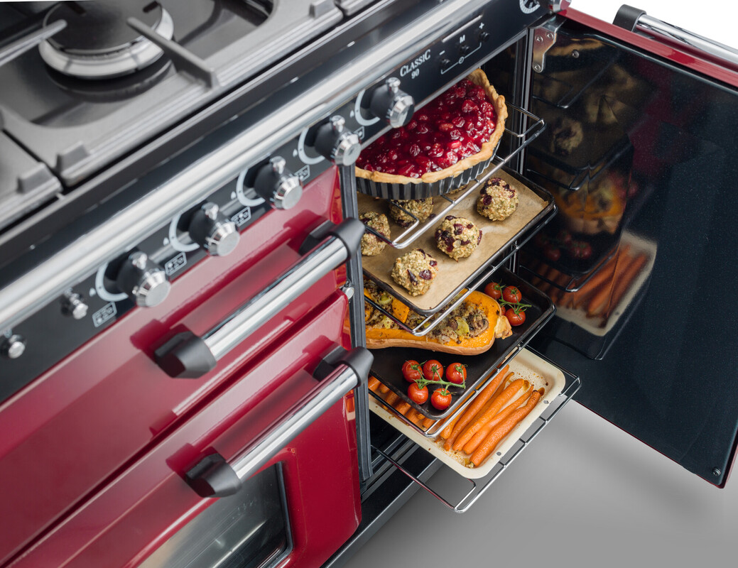 Ein geöffneter, moderner, roter Backofen mit verschiedenen Gerichten auf Backblechen: Ein Obstkuchen, Kekse, gefüllter Kürbis, Kirschtomaten und Möhren.