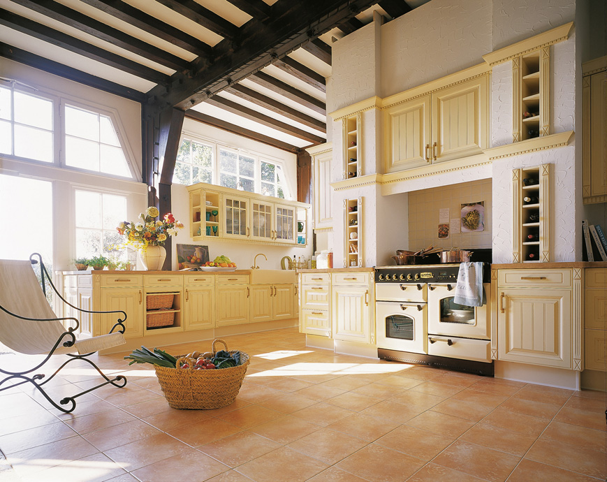 Lichtdurchflutete Landhausküche mit gelben Schränken, großem Herd und Korb mit frischem Gemüse auf dem Fliesenboden