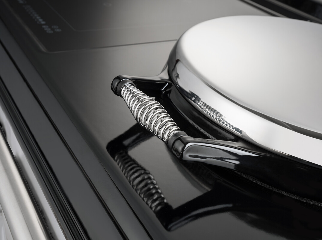 Nahaufnahme einer glänzenden, runden Metallabdeckung mit Feder-Griff auf einer schwarzen Herdplatte