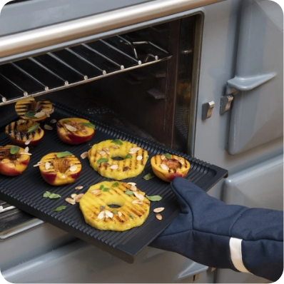 Eine Hand mit Ofenhandschuh holt eine Grillplatte mit gegrilltem Obst wie Ananasscheiben und Pfirsichen aus dem Ofen heraus.