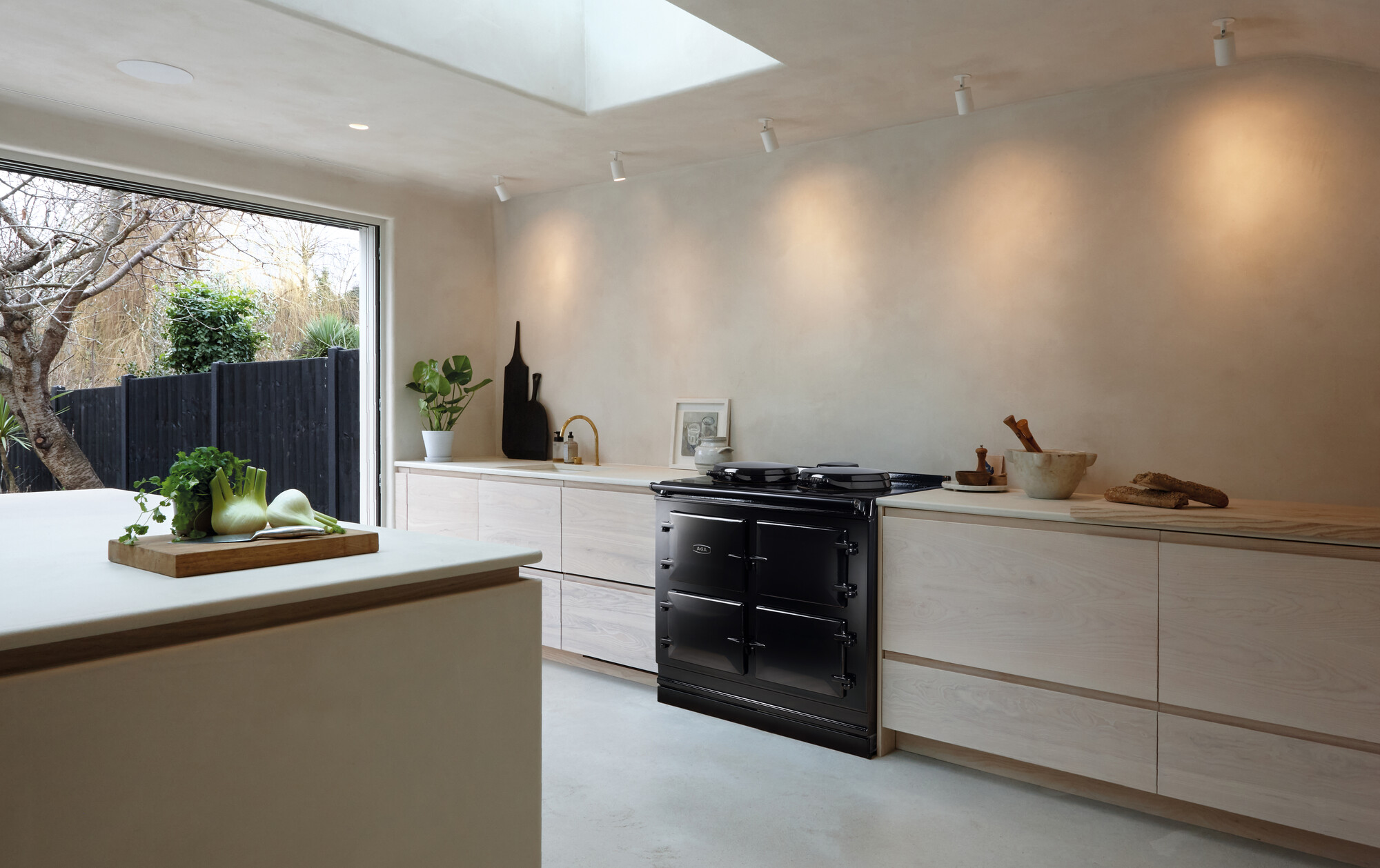 Minimalistisch eingerichtete Küche mit cremefarbenen Wänden, großem schwarzen Herd, hellen Holzschränken, Küchenutensilien und Ausblick in den Garten