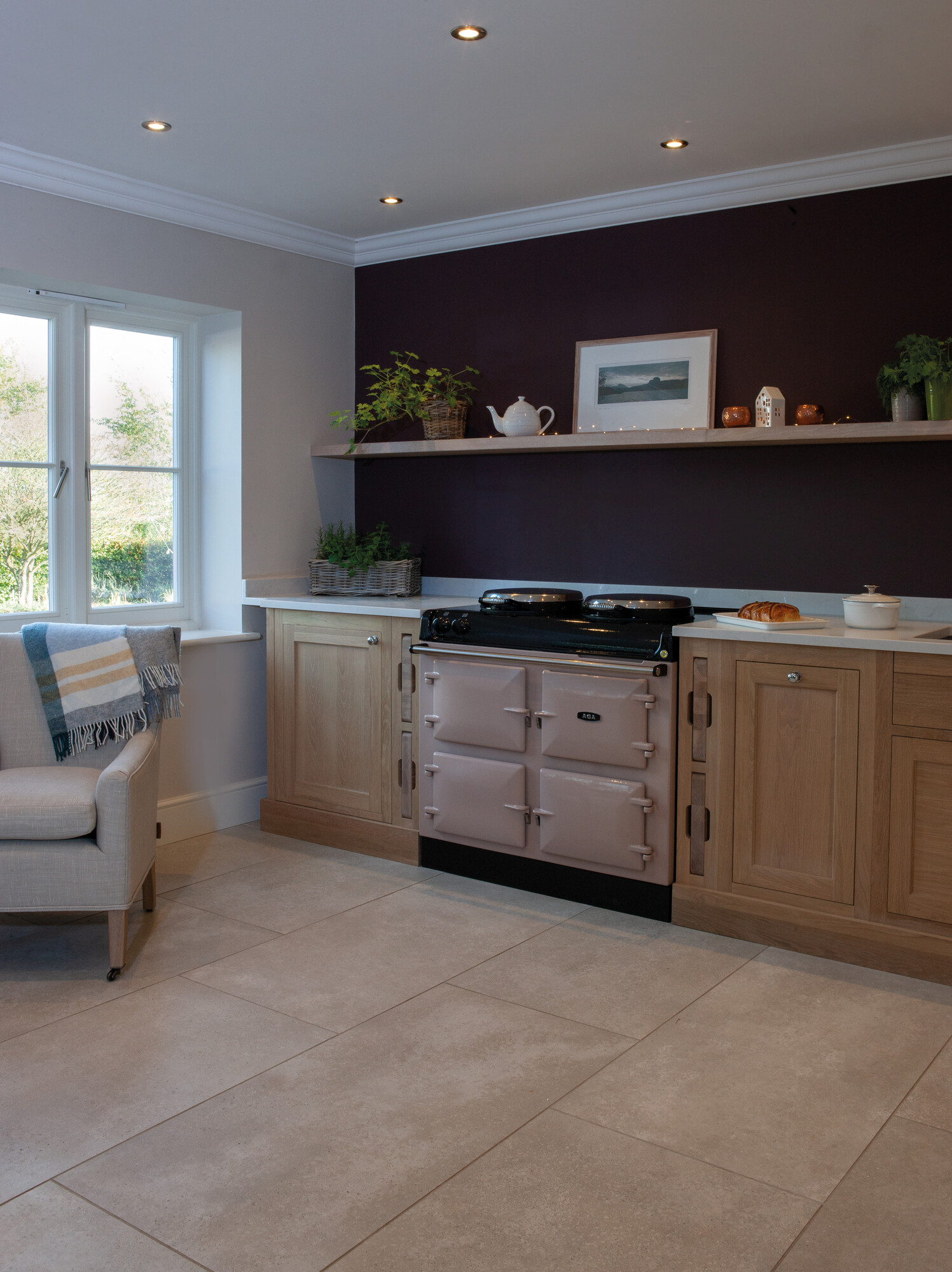 Gemütliche Küche mit cremefarbenem AGA-Herd, hellen Holzmöbeln, dunkler Wand, Fenster, Pflanzen und schlichtem Dekor